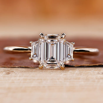 Lab created three stones engagement ring featuring an emerald cut center diamond with trapezoid side diamonds in a cathedral setting.