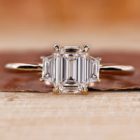 Lab created three stones engagement ring featuring an emerald cut center diamond with trapezoid side diamonds in a cathedral setting.