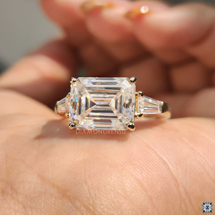 Three stone engagement ring featuring emerald cut moissanite and tapered baguette side stones.
