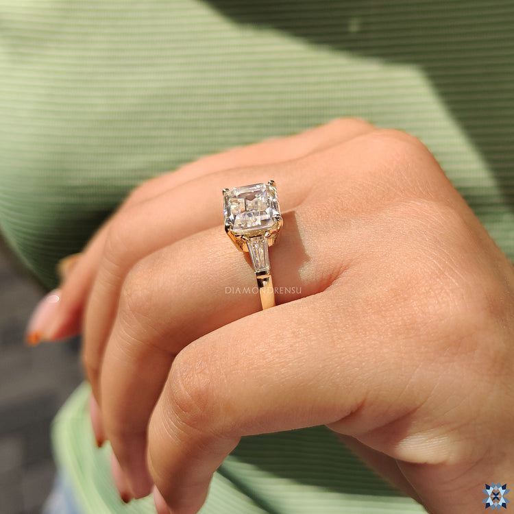 Four prong engagement ring with emerald cut moissanite and tapered baguette design.
